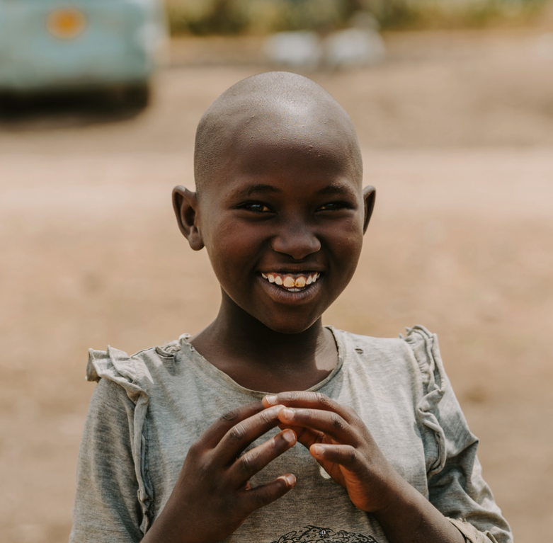A smiling young girl with a shaved head, wearing a light gray shirt with ruffled sleeves, standing outdoors on a dirt surface.