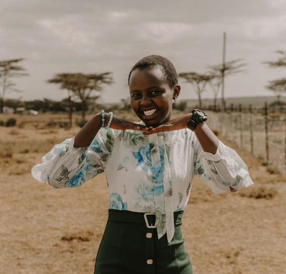 Young girl with short hair smiling and holding her hands under her chin outdoors in a rural setting.