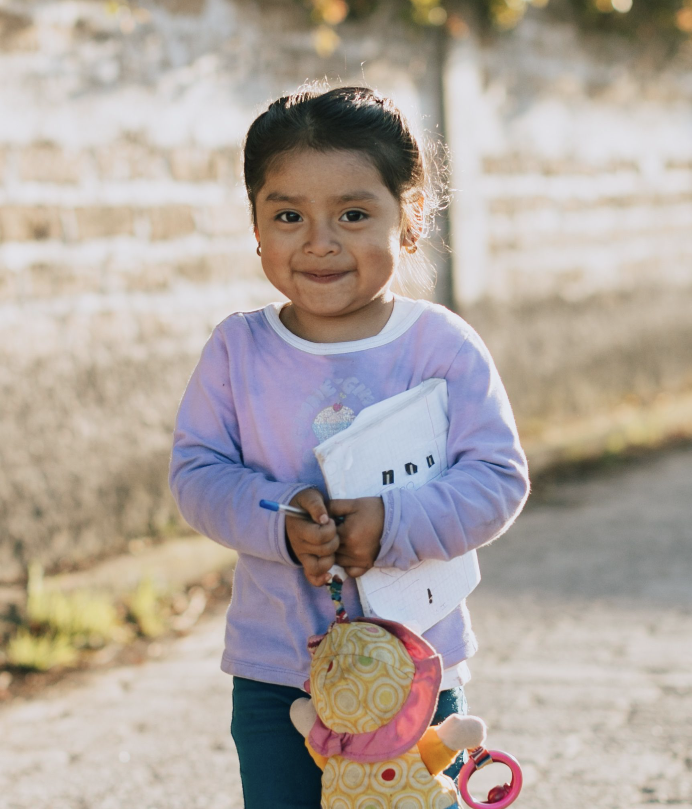 Young girl smiling outdoors, holding a notebook and a soft toy, with a blurred background of trees and a brick wall, bathed in warm sunlight.