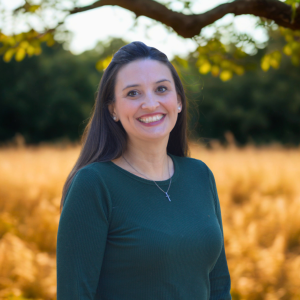 A woman with long dark hair smiling outdoors in a field with trees in the background.
