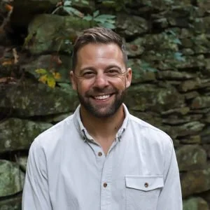 A smiling man with short hair and a beard, wearing a white button-up shirt, standing outdoors in front of a stone wall with greenery.