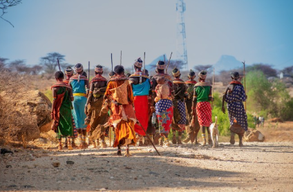 Group of people walking together outdoors in a dry, rural area, some carrying walking sticks, with sparse vegetation and a clear sky.