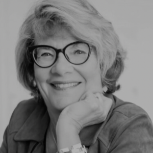 A smiling woman with glasses and short wavy hair, resting her chin on her hand.