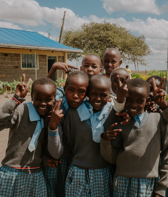 Group of six smiling school children in uniform making peace signs outdoors in front of a school building and tree.