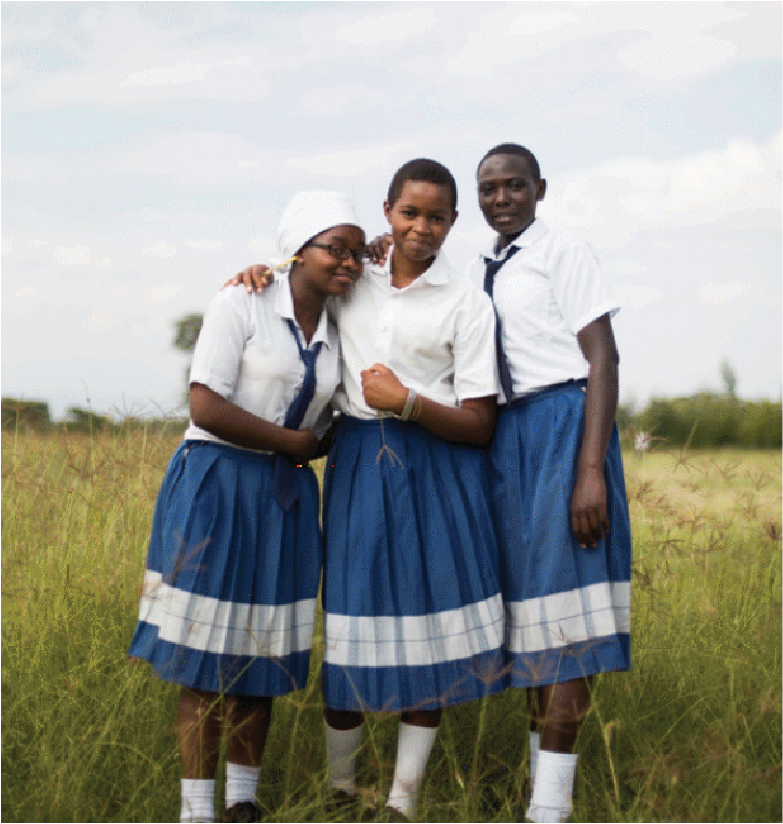 Three students in school uniforms standing together outdoors in a grassy field, smiling and posing for the photo.