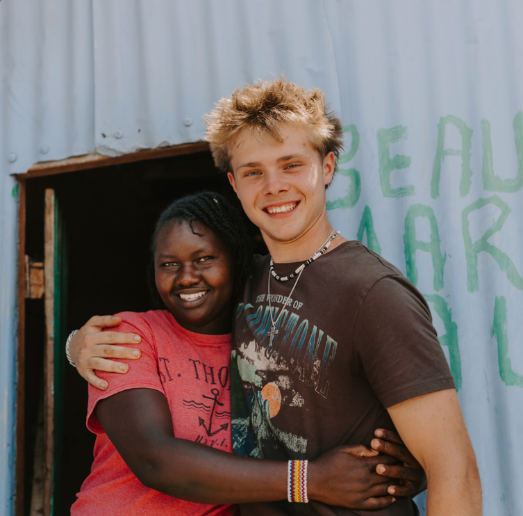 Two people hugging in front of a blue wall with green lettering, smiling at the camera.
