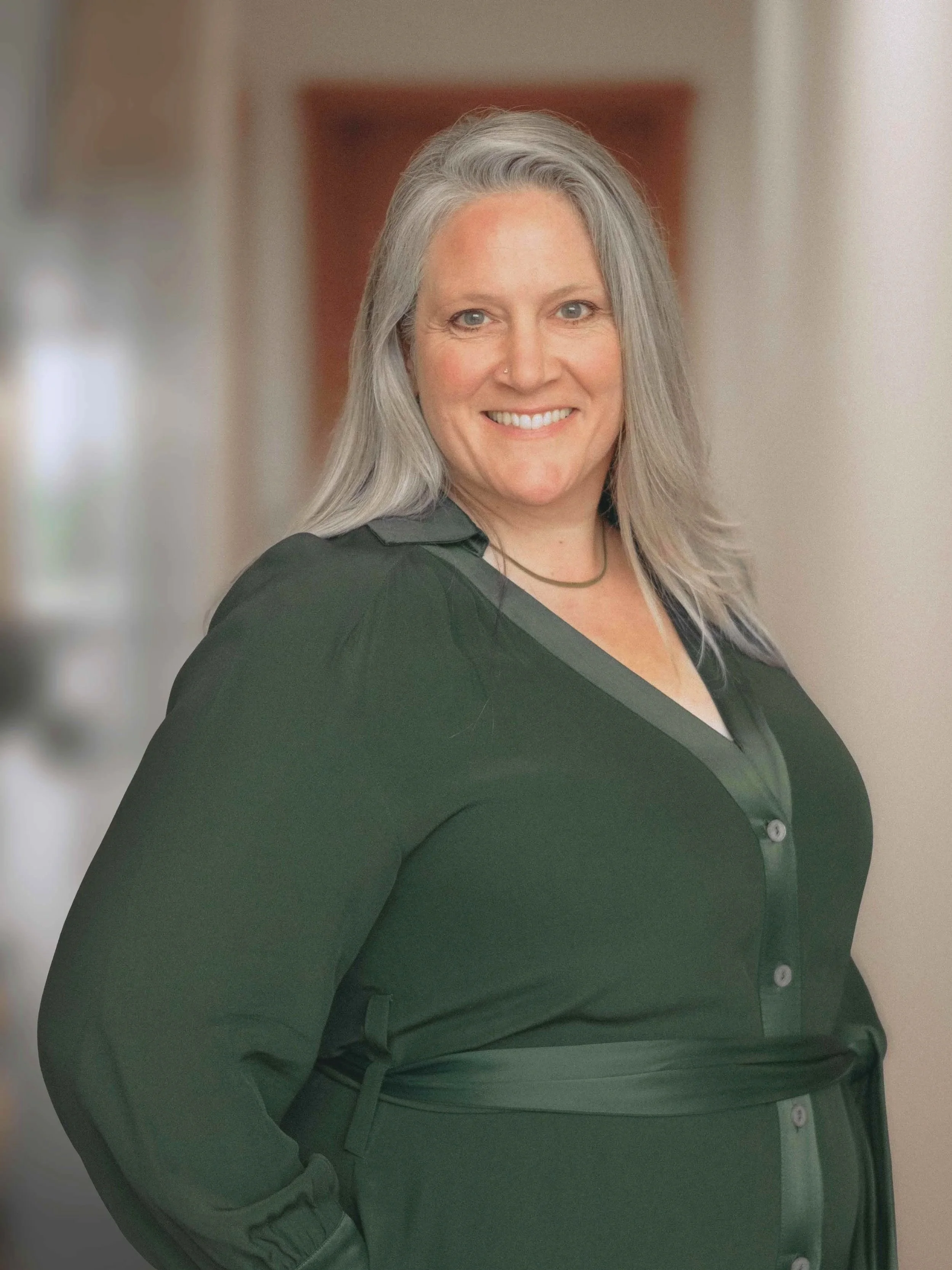 A smiling woman with shoulder-length gray hair wearing a dark green blouse, standing indoors with a blurred background.