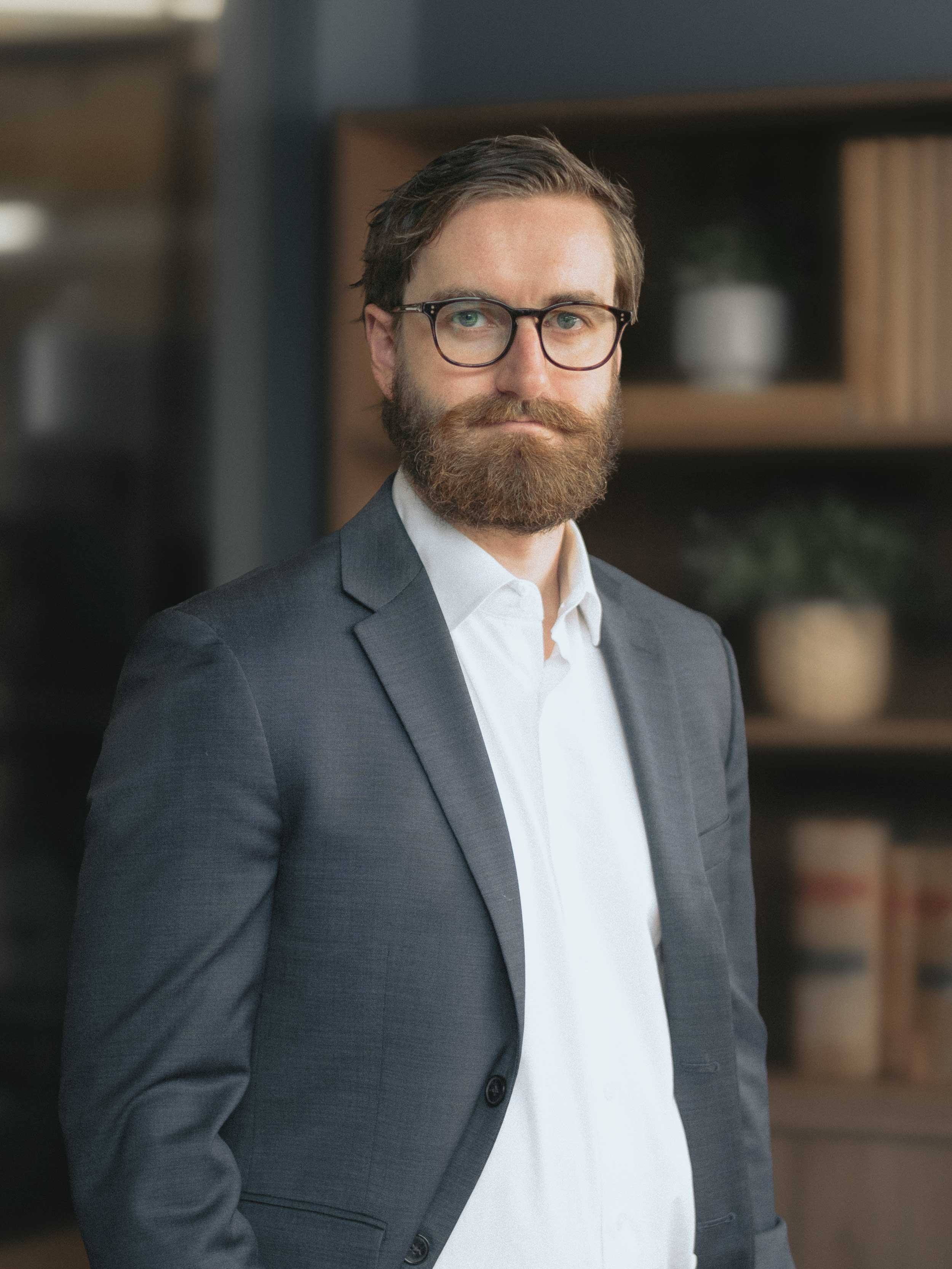 A man with glasses, a full beard, and brown hair, wearing a gray suit and white shirt, standing indoors with blurred background of shelves and plants.