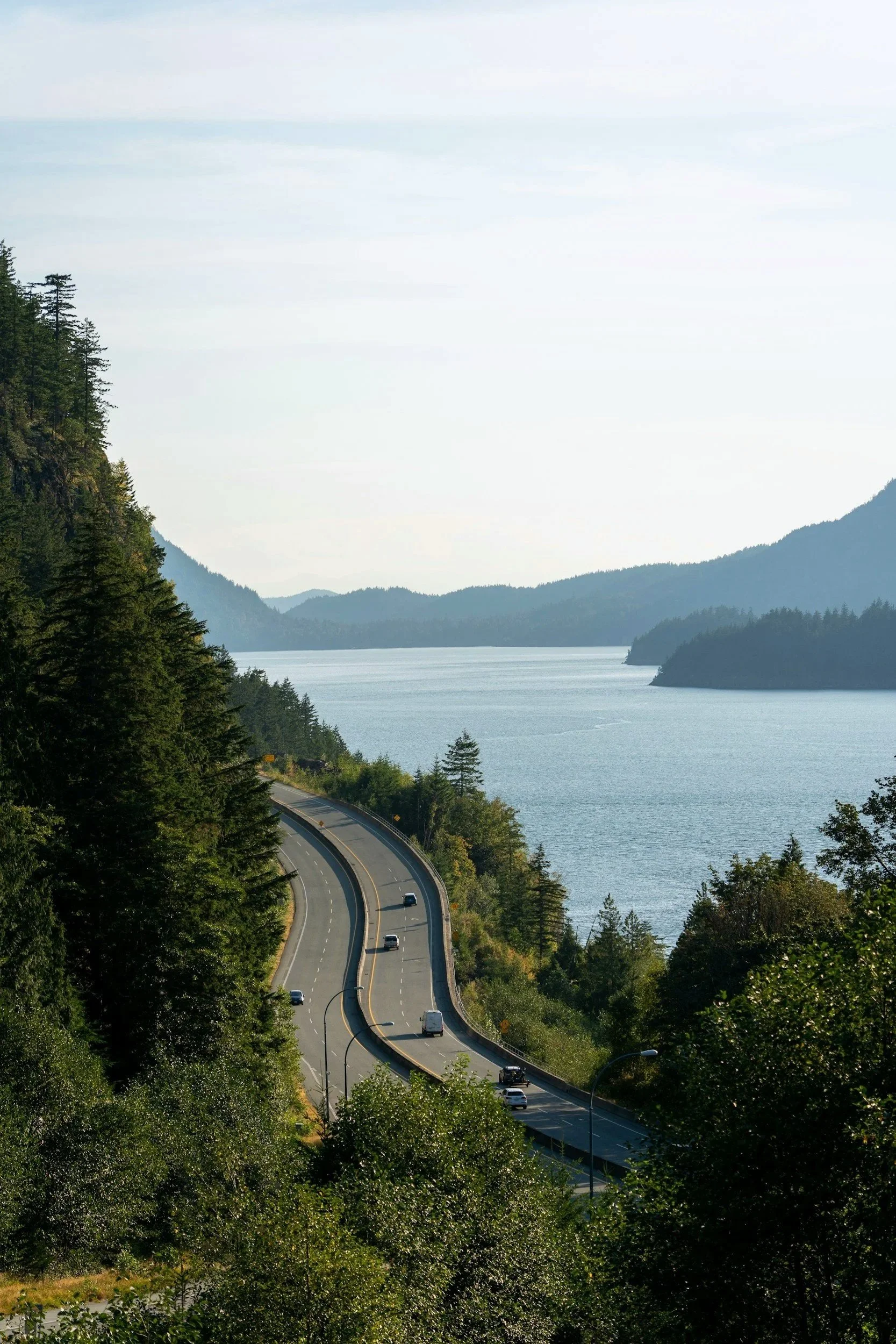 A winding road runs along a wooded hillside overlooking a large body of water, with mountains in the background and a clear sky overhead.