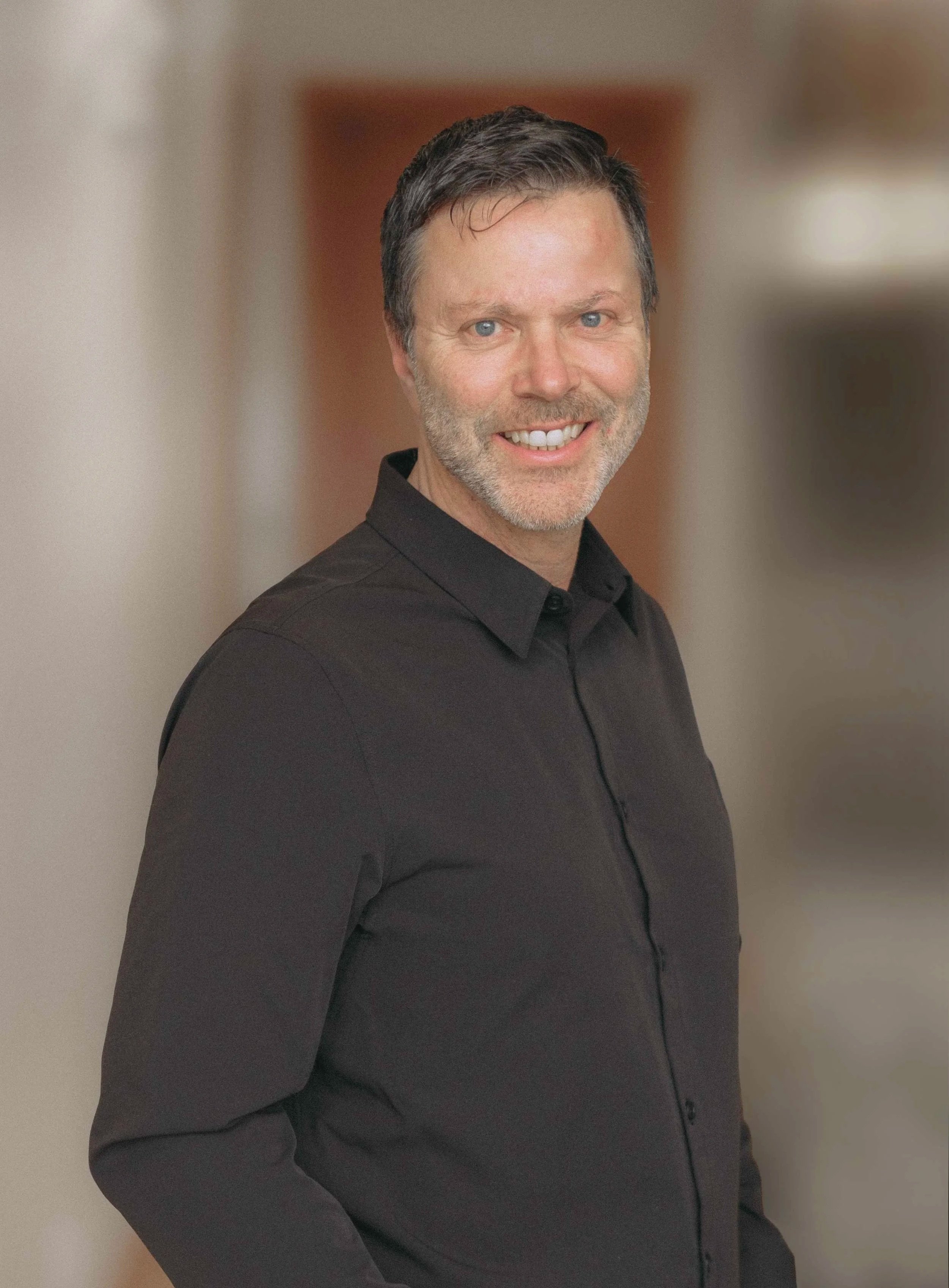 A smiling man with dark hair and a beard, wearing a black shirt, standing in an indoor setting with a blurred background.