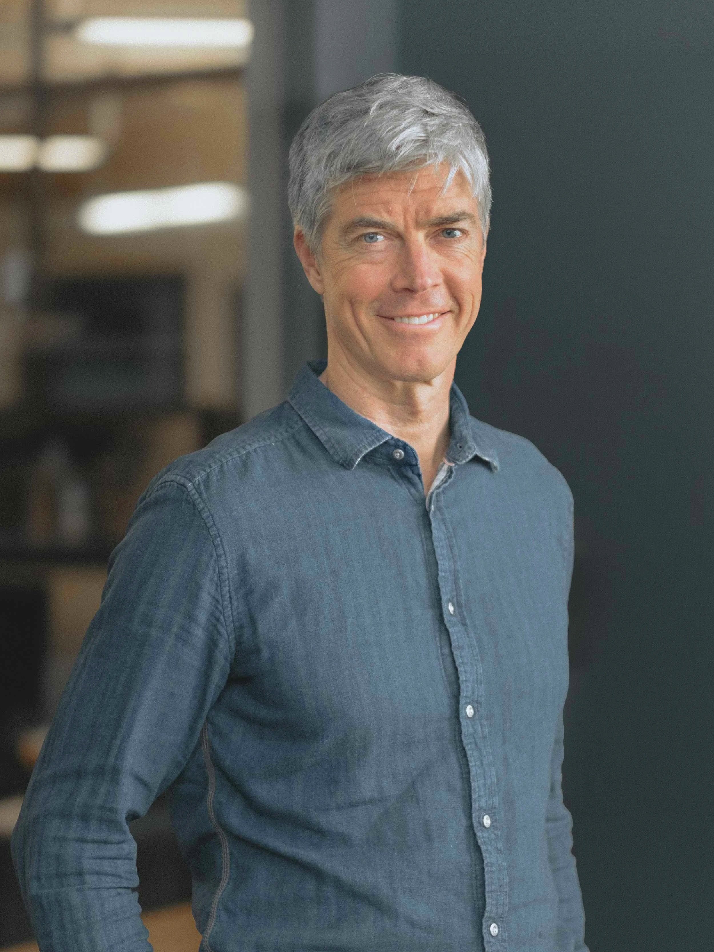 Middle-aged man with gray hair smiling, wearing a denim shirt, standing indoors.