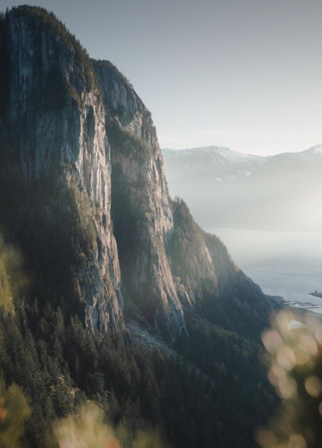 A mountain with steep cliffs and forested slopes overlooking a body of water, with distant mountain ranges in the background.