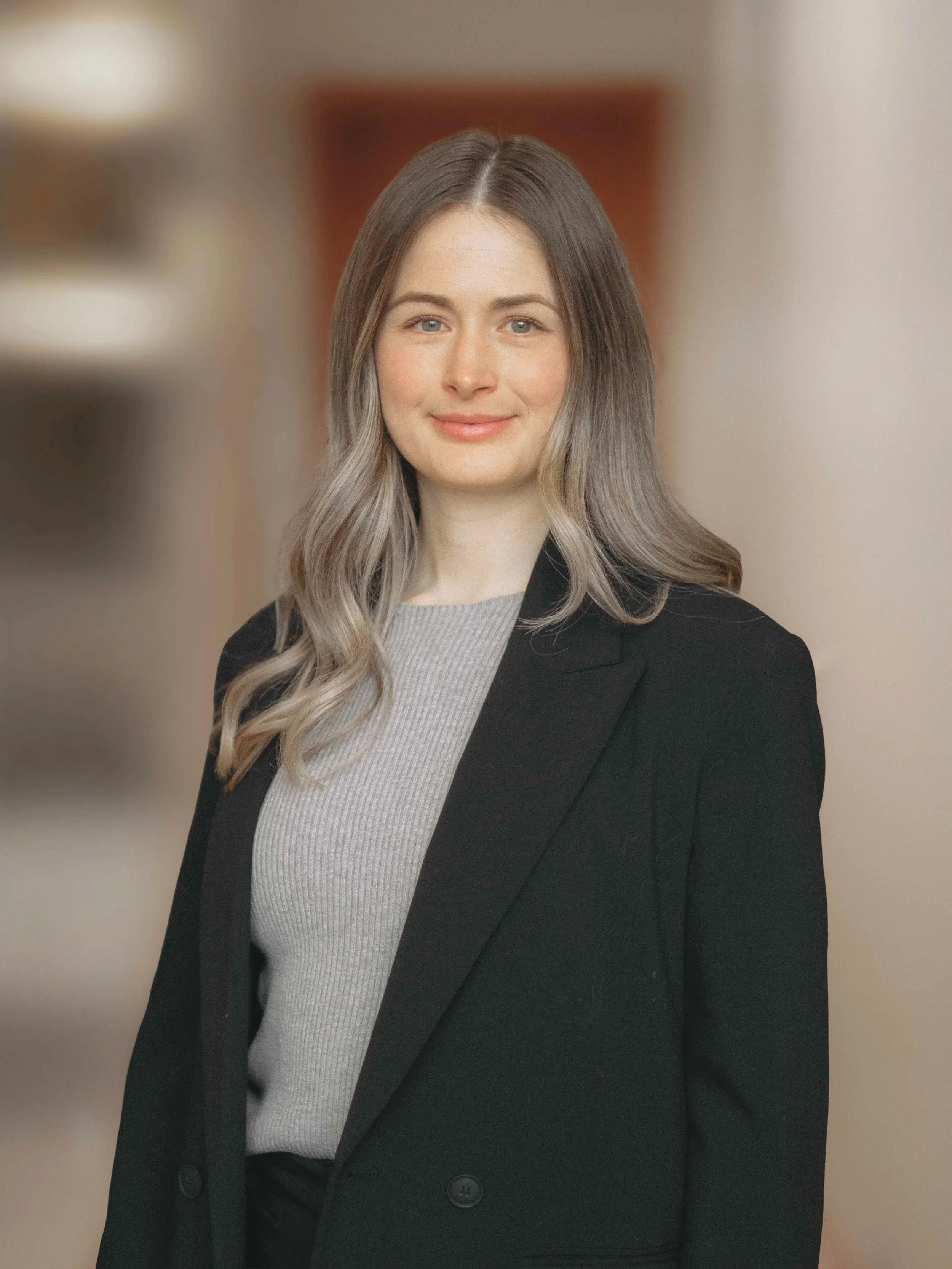 A young woman with long, wavy blonde hair and blue eyes, wearing a gray top and a black blazer, smiling against a blurred indoor background.
