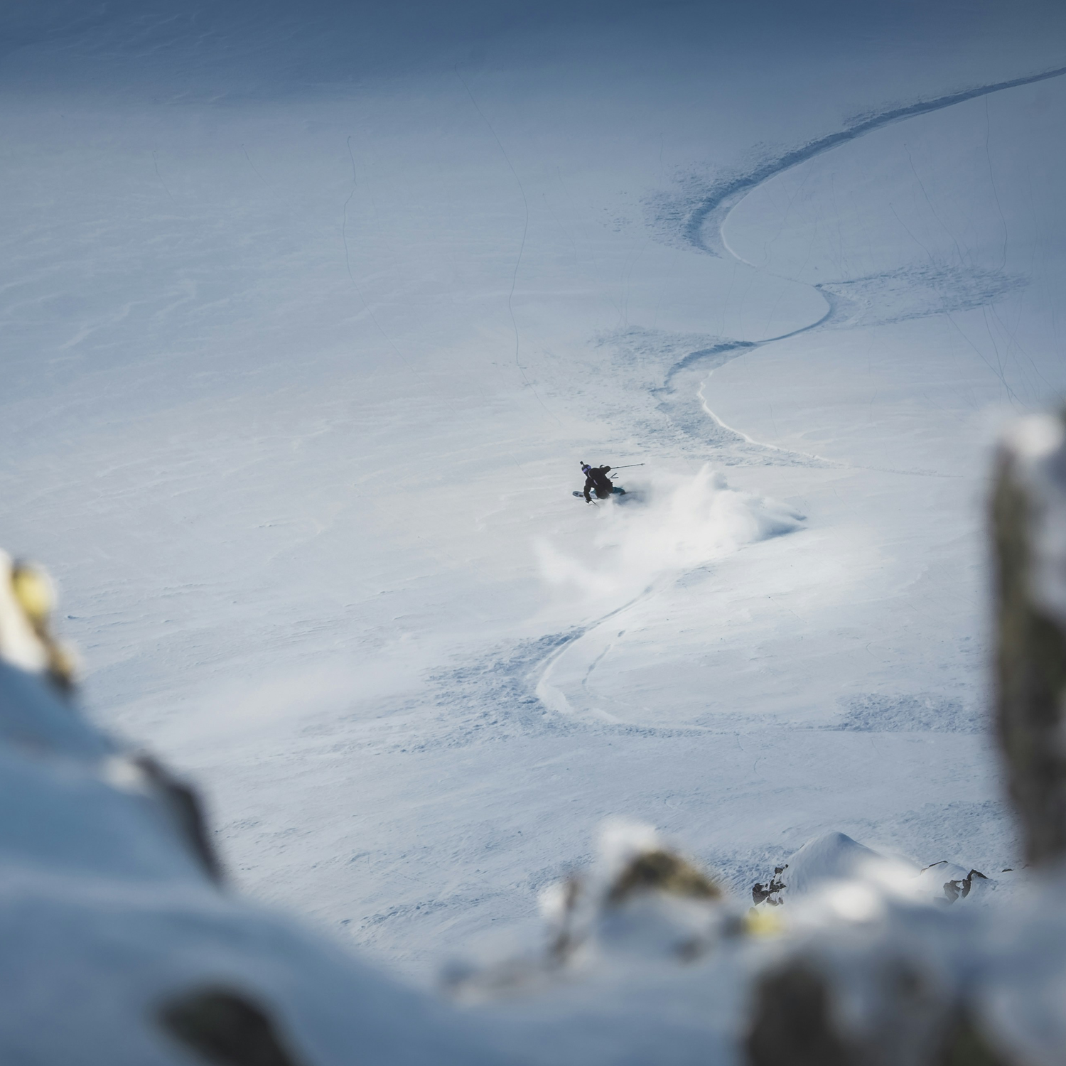 A person snowboarding on a snowy mountain slope, leaving a trail in the snow, with snow-covered rocks in the foreground.