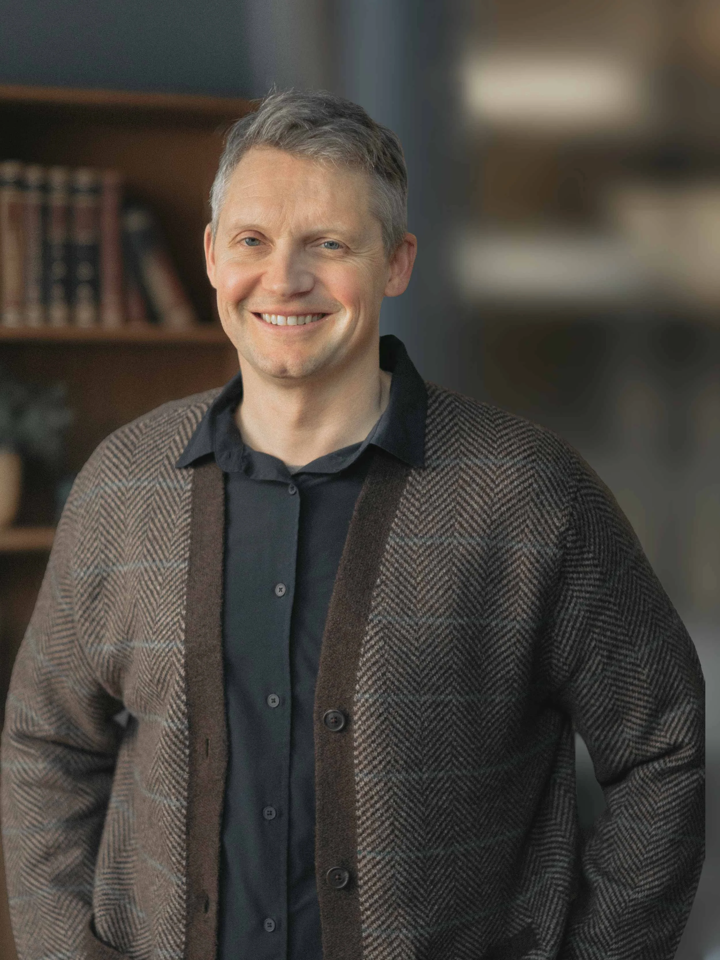 A smiling middle-aged man with gray hair, wearing a black shirt and a brown, herringbone-patterned cardigan, standing indoors with bookshelves in the background.