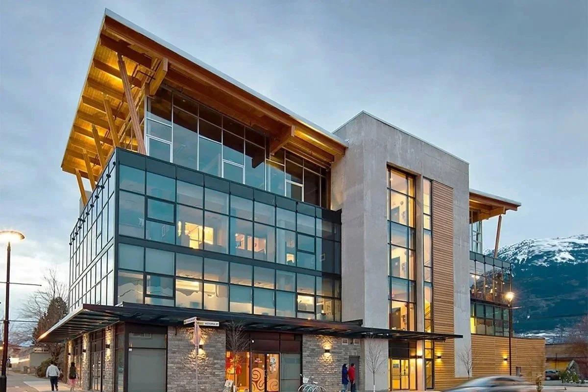 Modern multi-story building with glass windows and wooden accents, situated near snow-capped mountains, with people walking outside during evening.