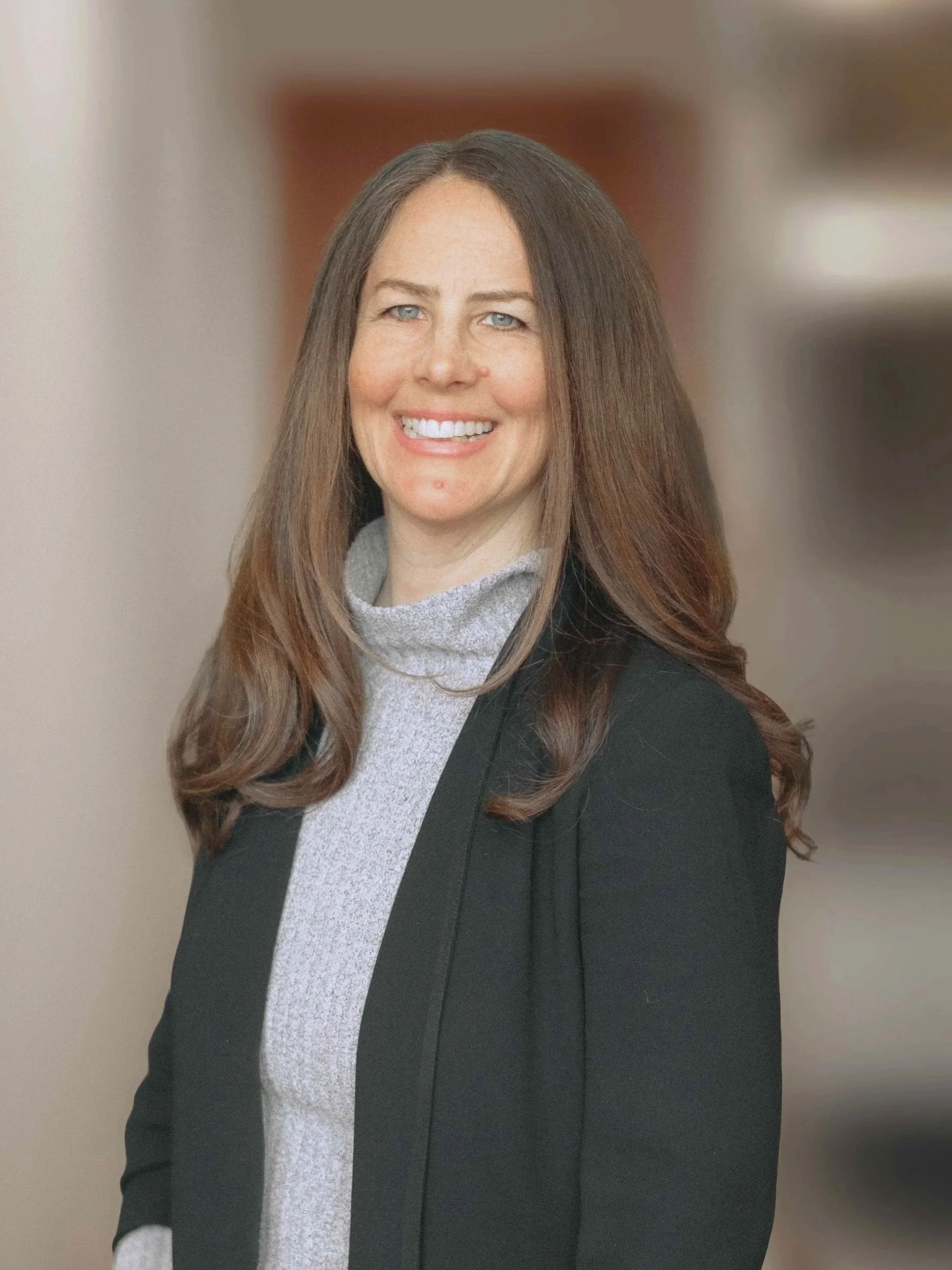 A smiling woman with long brown hair, wearing a black blazer and light gray turtleneck, standing indoors with a blurred background.