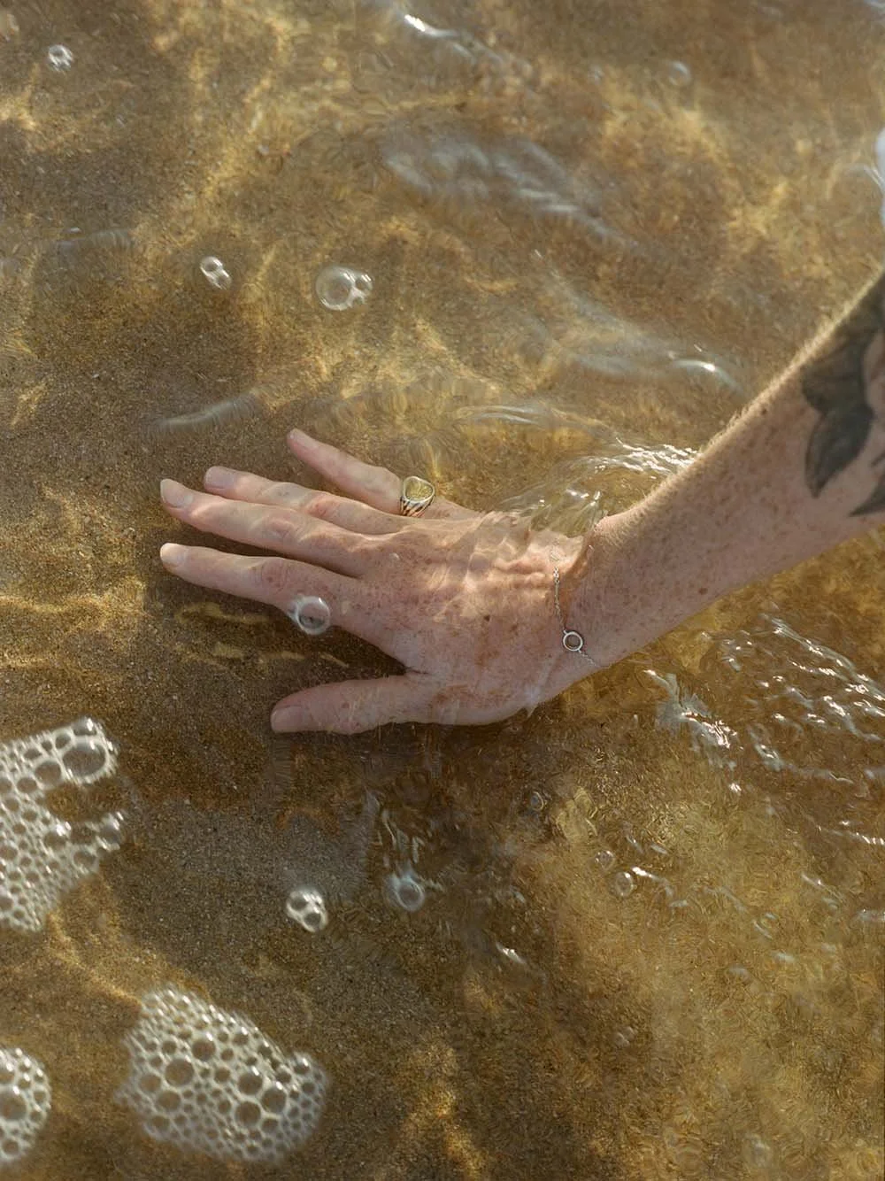 A person's hand resting on sandy riverbed with clear water flowing over it.