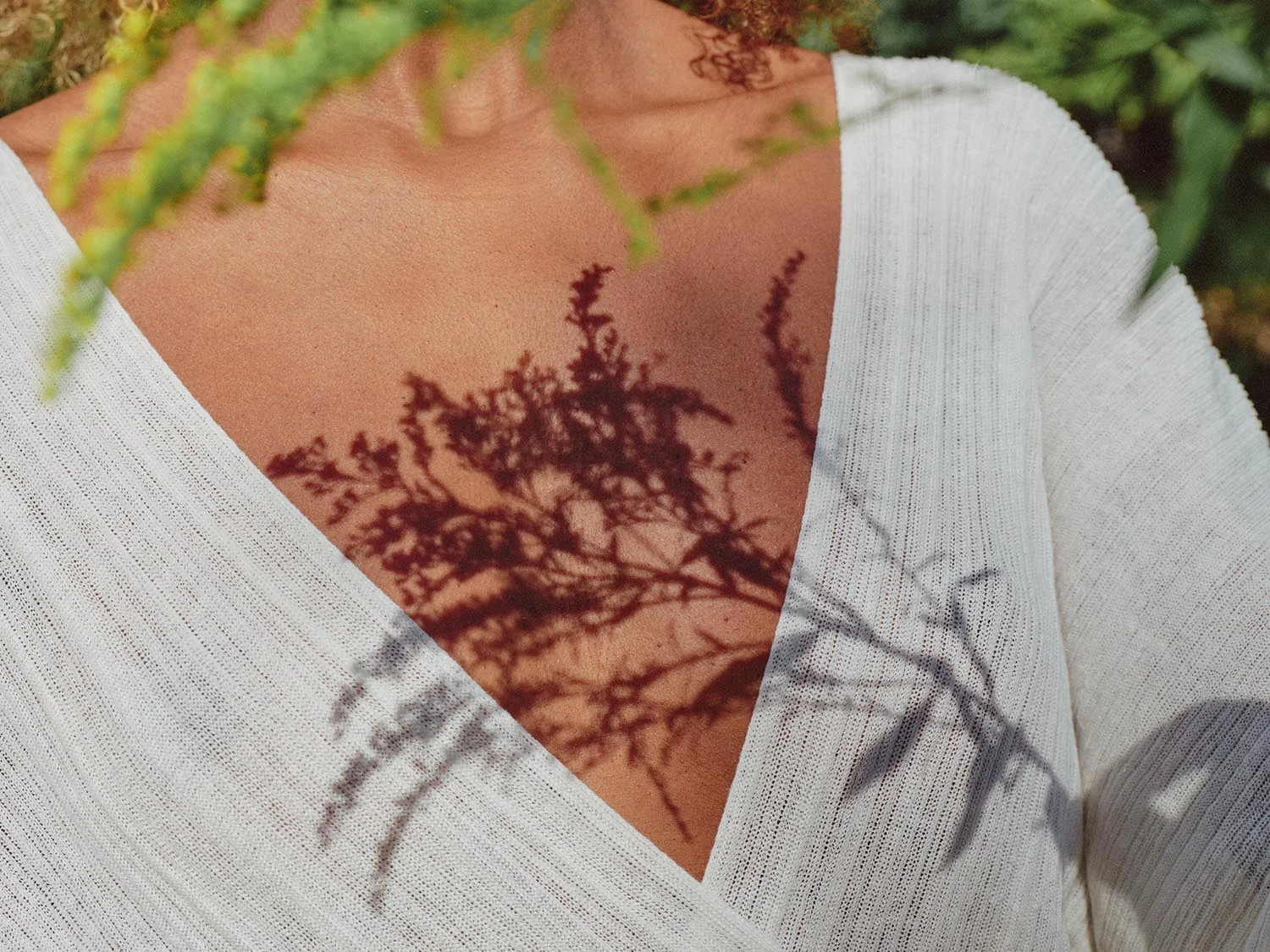 A shadow of a leafy plant cast on a woman's chest and dress, with the woman wearing a white V-neck top in an outdoor setting.