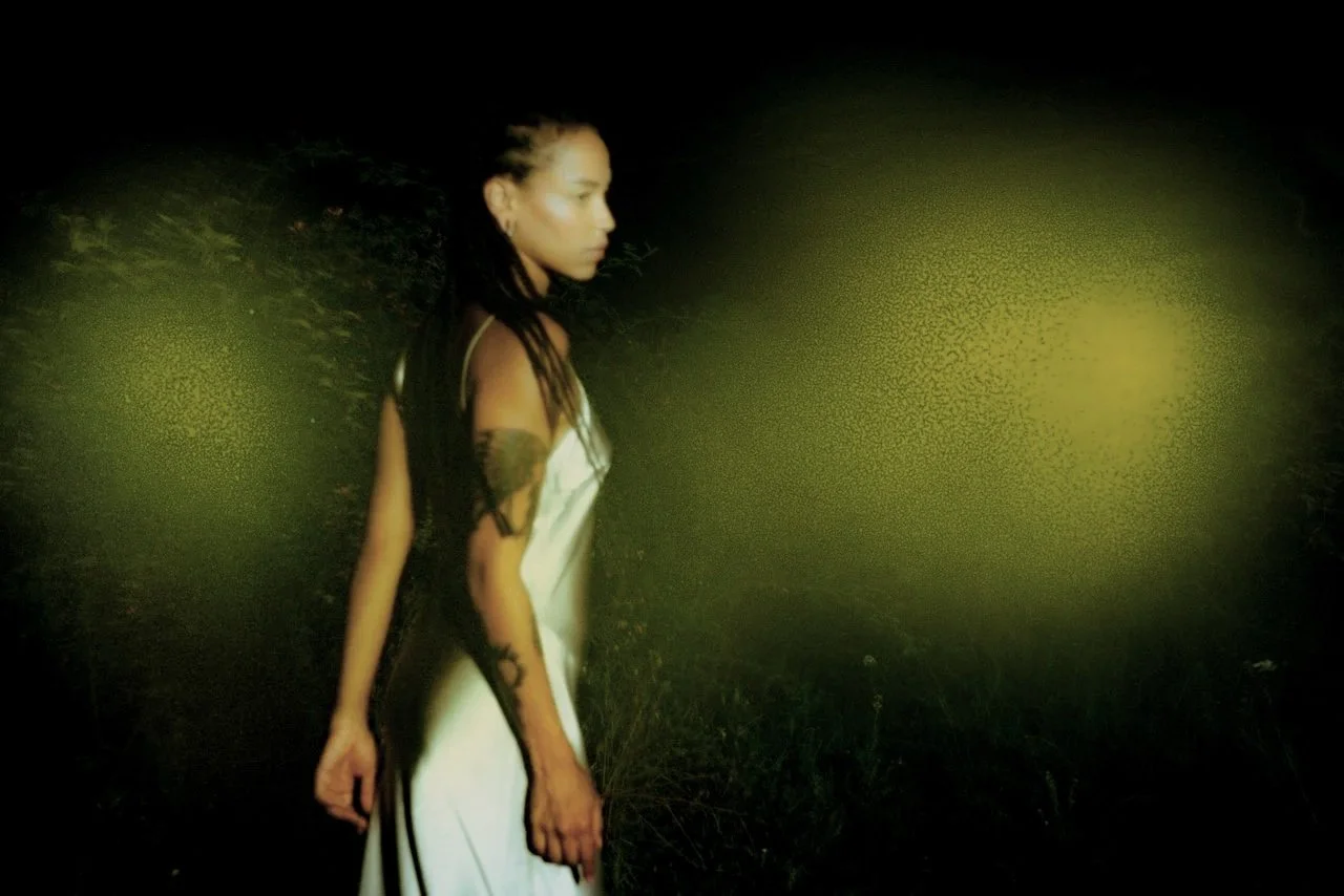 A woman with long dark hair and tattoos on her arm, wearing a white dress, standing outdoors at night with blurred greenish lights in the background.