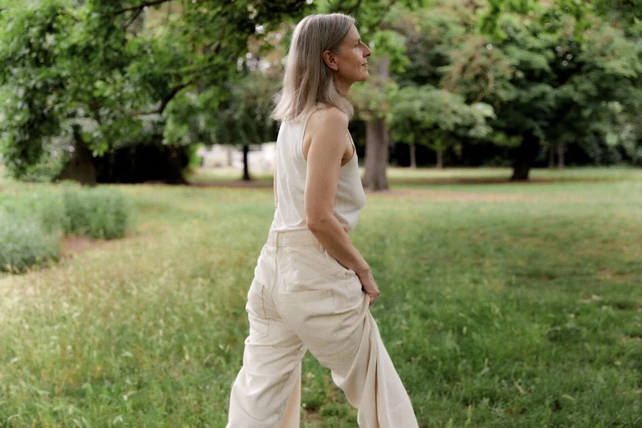A woman with shoulder-length gray hair walking in a park with green trees and grass, wearing a sleeveless beige top and baggy beige pants.