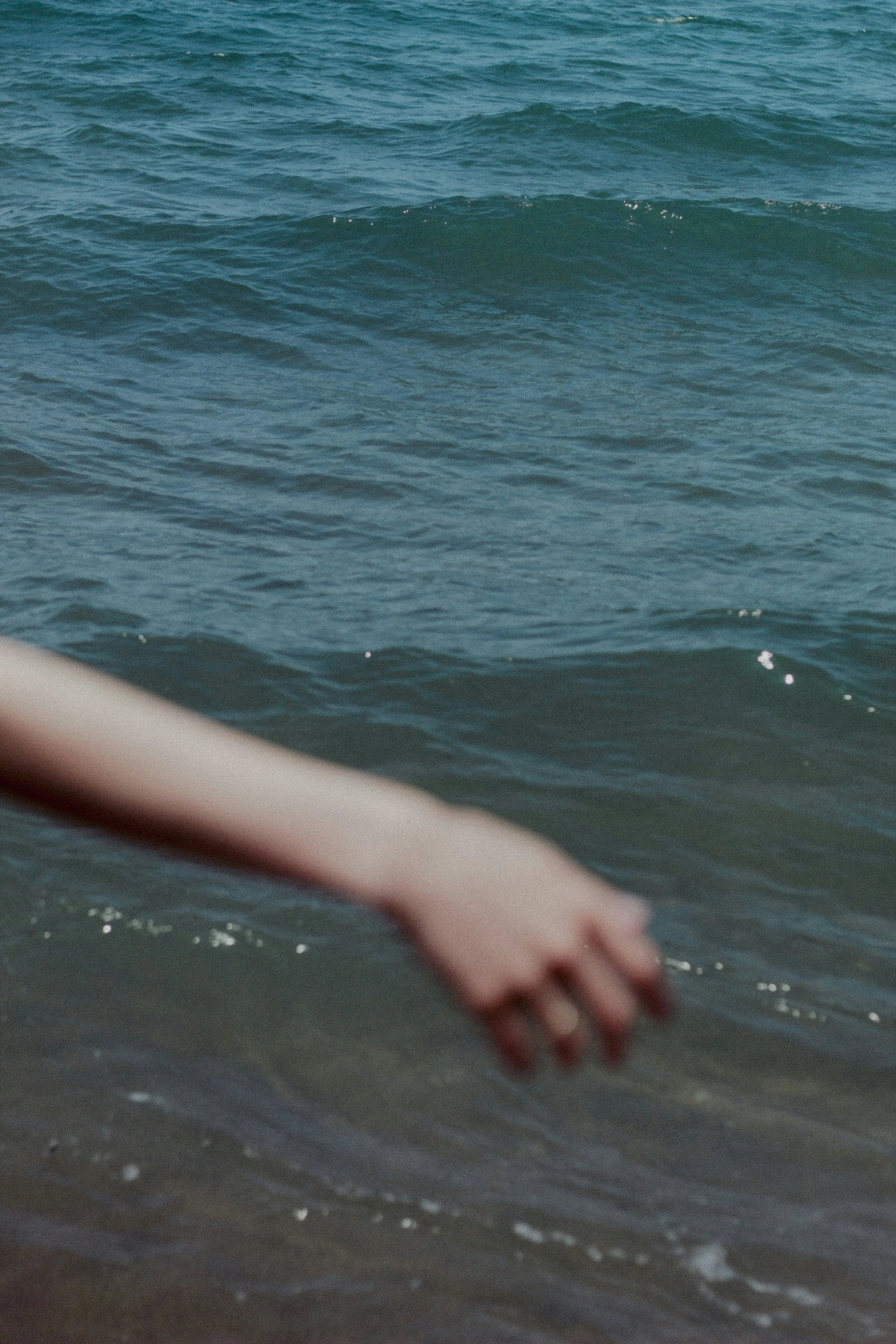 A person’s arm and hand reaching into the water at the beach with ocean waves in the background.