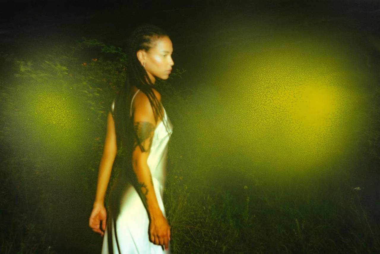 A woman with long dark hair, wearing a white dress with black lace details, standing outdoors at night with a greenish-yellow light creating an abstract glow in the background.