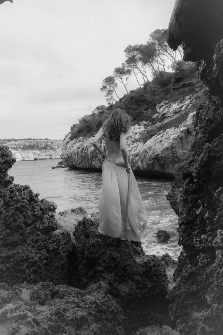 A woman with long hair standing on rocks near the water, wearing a flowing skirt, with trees and cliffs in the background, in black and white.