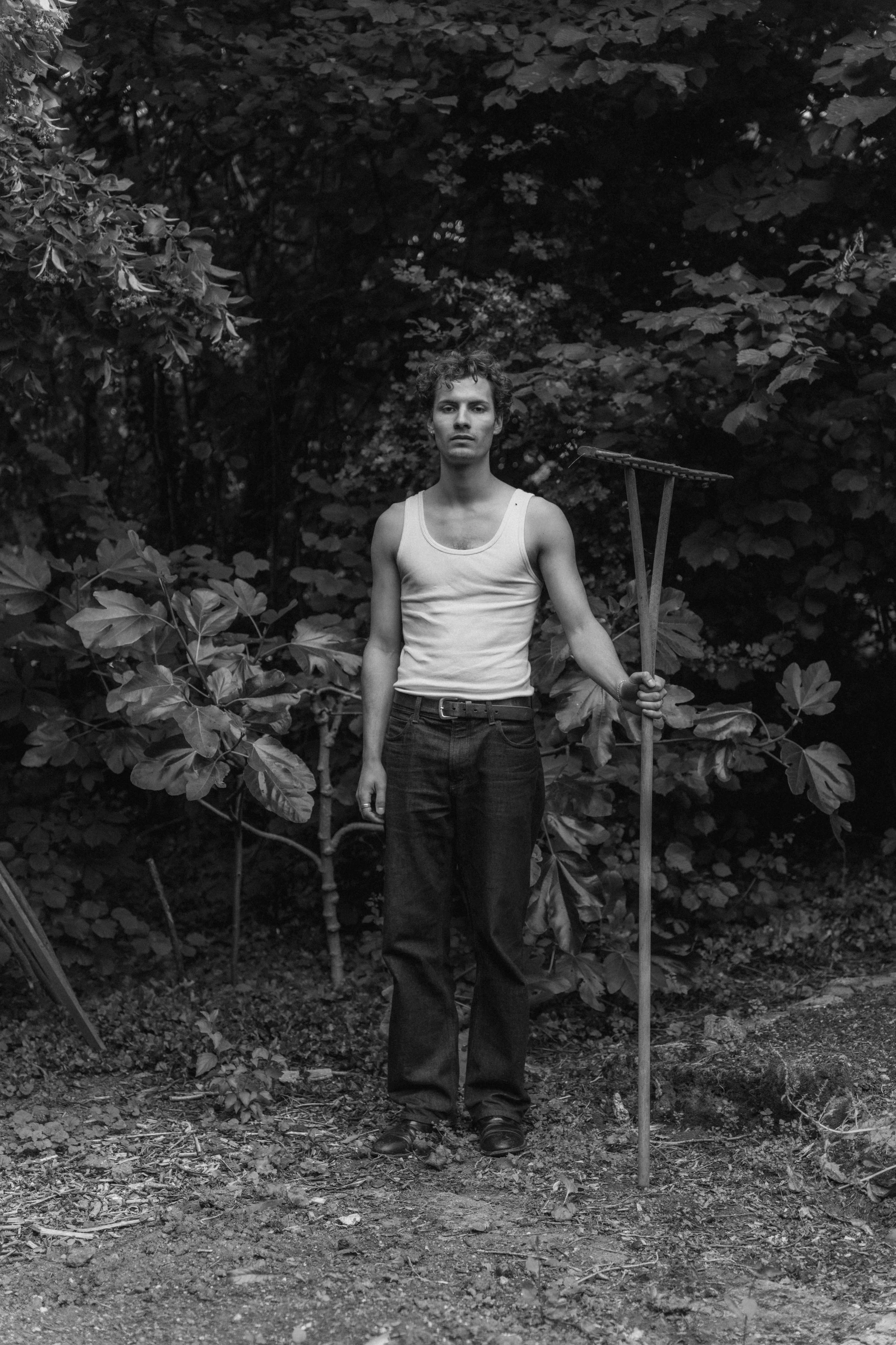 A young man standing in a forested area, holding a rake, wearing a tank top and jeans, with a serious expression.
