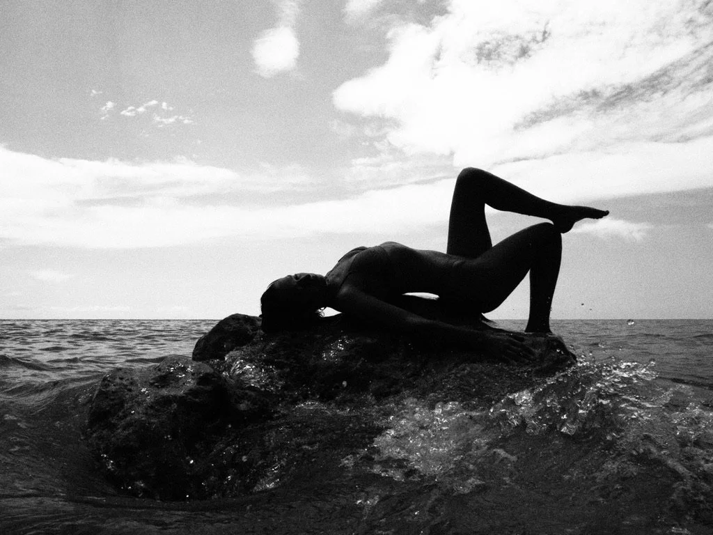 Silhouette of a woman lying on a rock in the water, with one leg bent and the other extended, against a cloudy sky background.