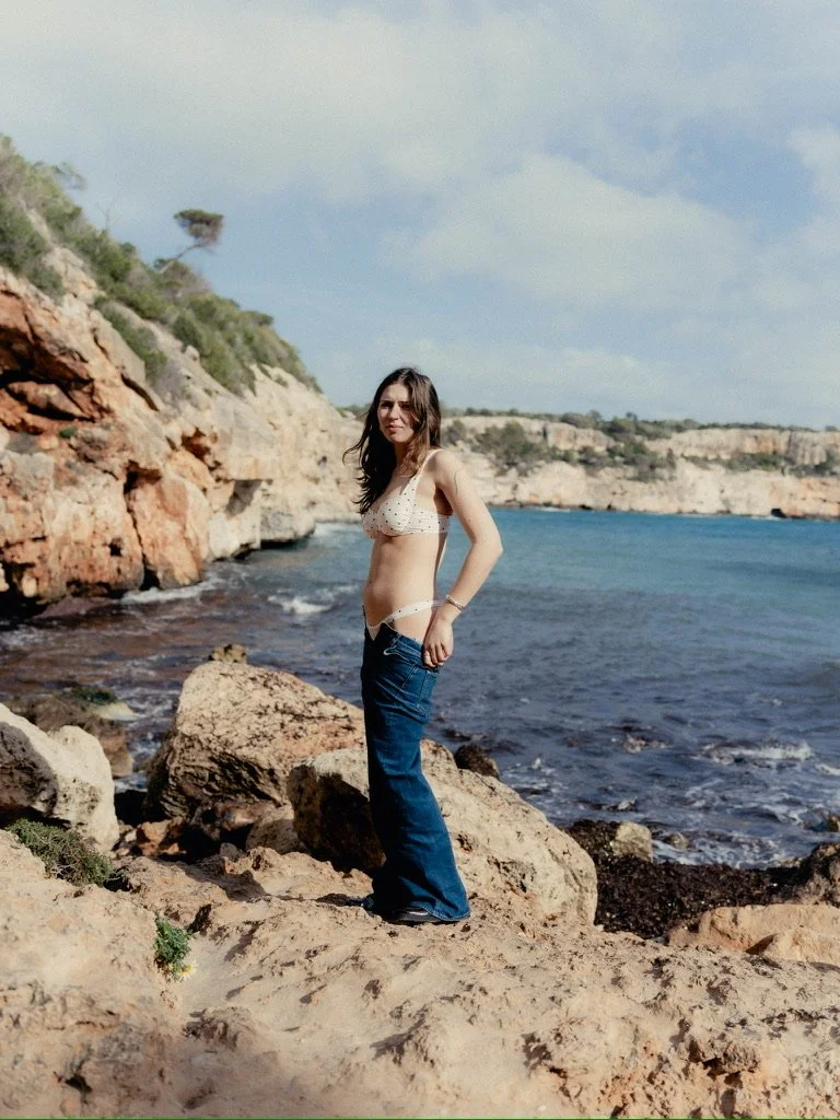 A woman standing on rocks by the ocean, wearing a bikini top and jeans, with cliffs and water in the background under a partly cloudy sky.
