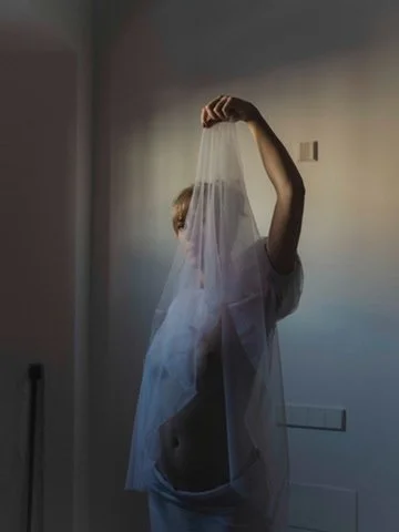 A woman in a sheer veil holding it up above her head, with a tattooed stomach visible, sitting indoors against a plain wall.