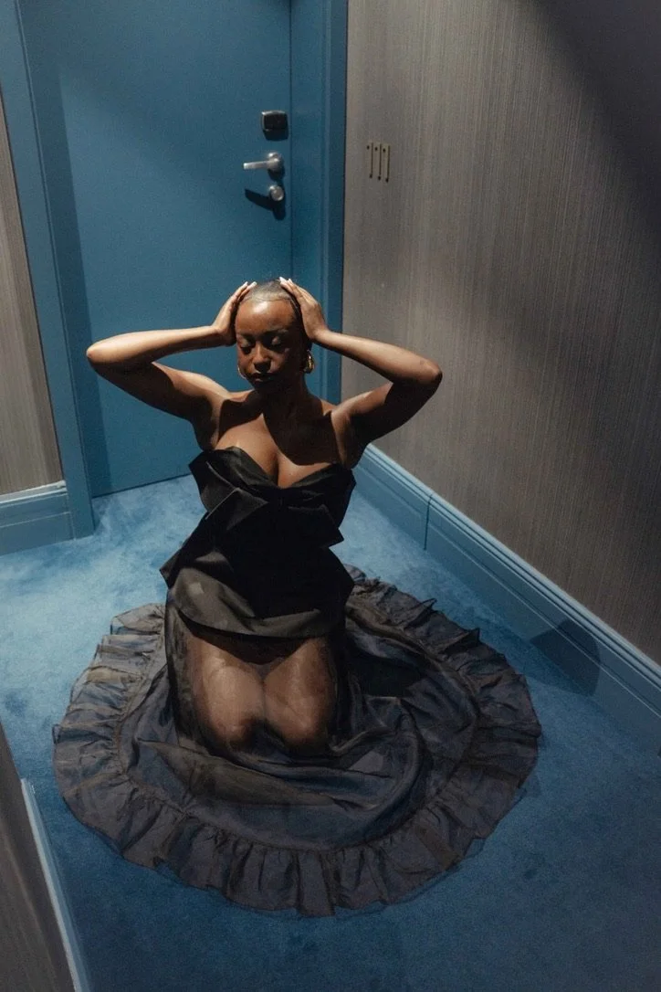 A woman kneels on a ruffled black dress on the floor in a small room, with her hands on her head and eyes closed.