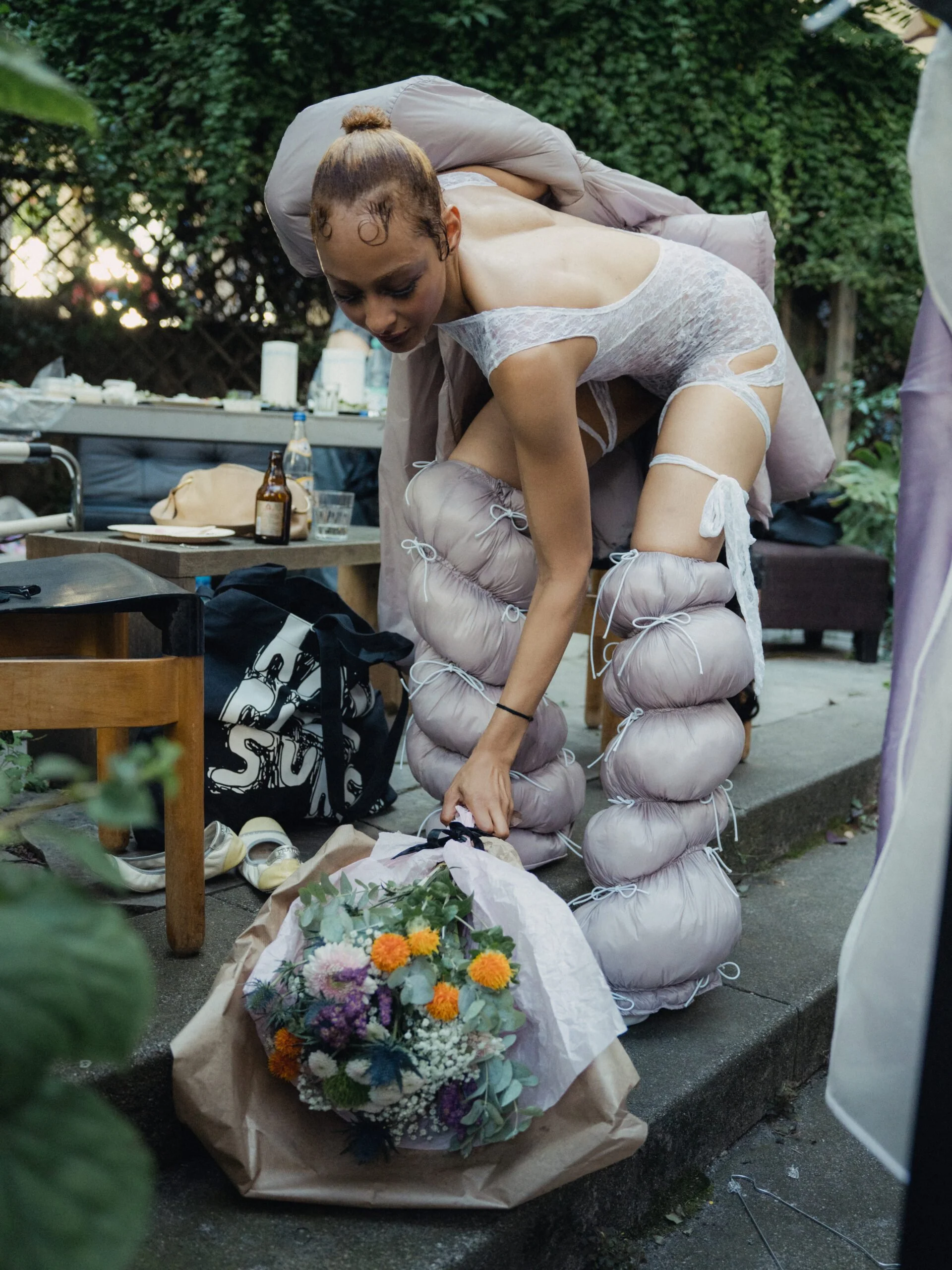 A woman dressed in a white lace dress and beige padded trousers, with her hair in a bun, is bowing her head at a memorial or funeral setting. There are flowers on the ground, a table with drinks and personal items, and a green leafy background.