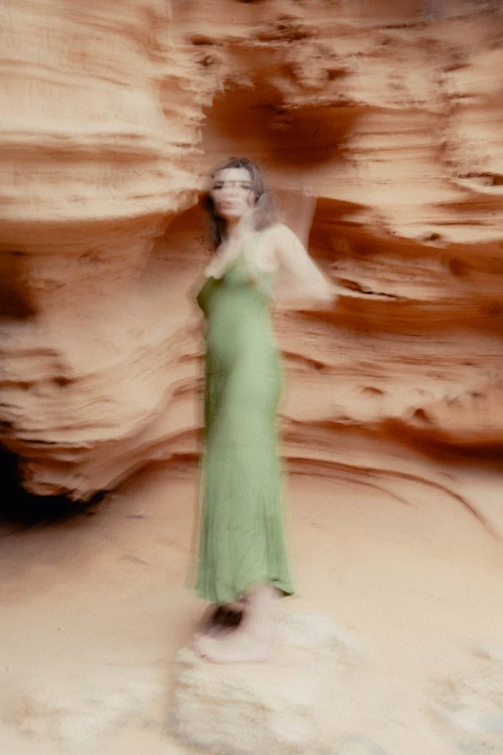 A woman standing on a rock in a desert with red sandstone formations, wearing a long green dress and glasses.