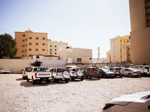 Parking lot with various cars and pickup trucks, with buildings and a minaret in the background under a clear sky.