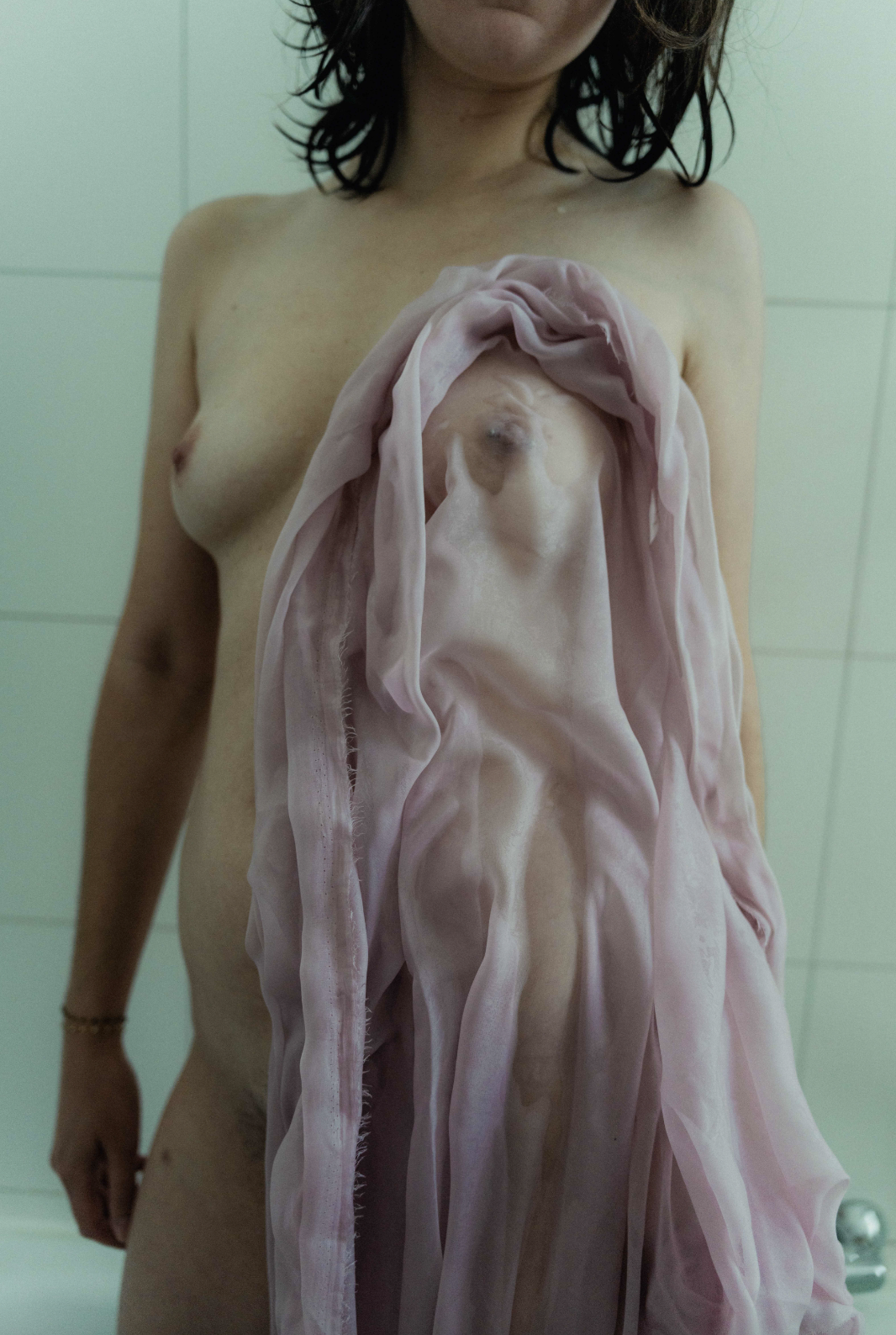 A woman with wet hair is standing topless with a pink fabric covering part of her body, in front of a pale wall, in a setting that appears to be a bathroom or studio.