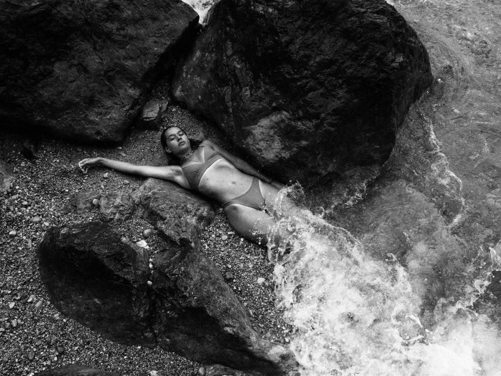 A woman in a bikini lying on a bed of rocks between large rocks near water, with one arm stretched out and water flowing around her.