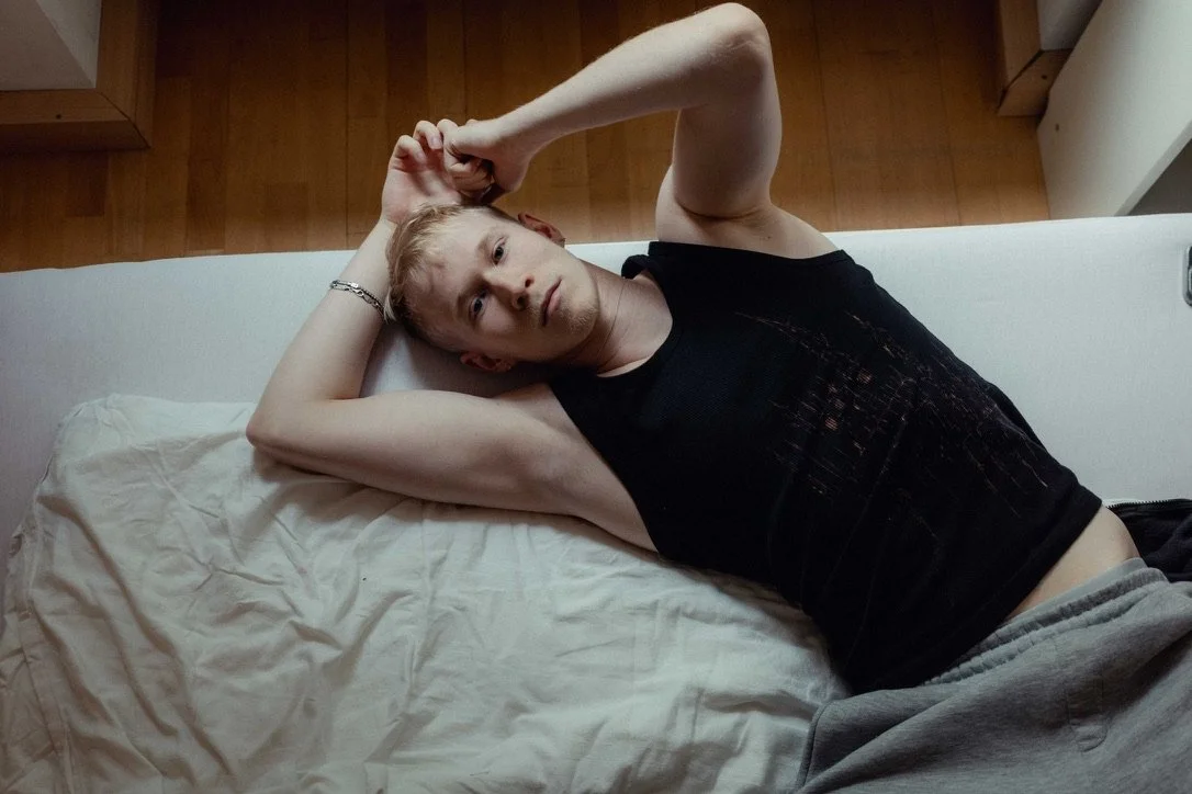 Young man lying on a bed, wearing a black tank top and gray sweatpants, looking at the camera with a neutral expression, with one arm resting behind his head and the other bent upward.