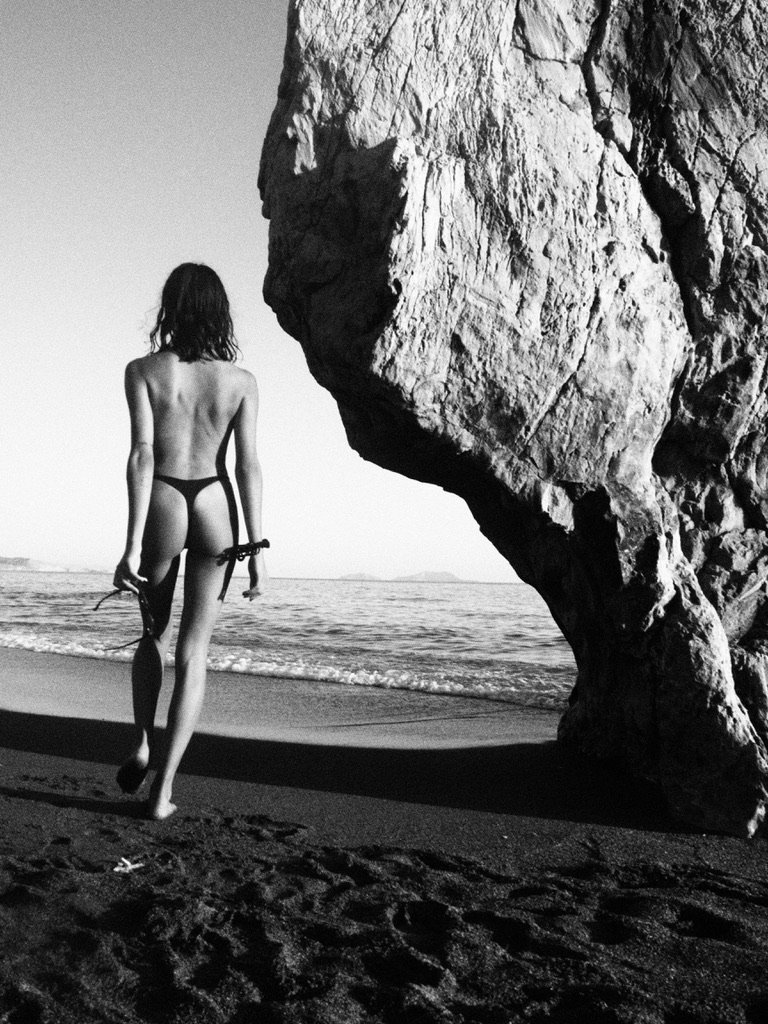 Woman walking on beach near large rock formation, holding a towel and swimwear, black and white photo.