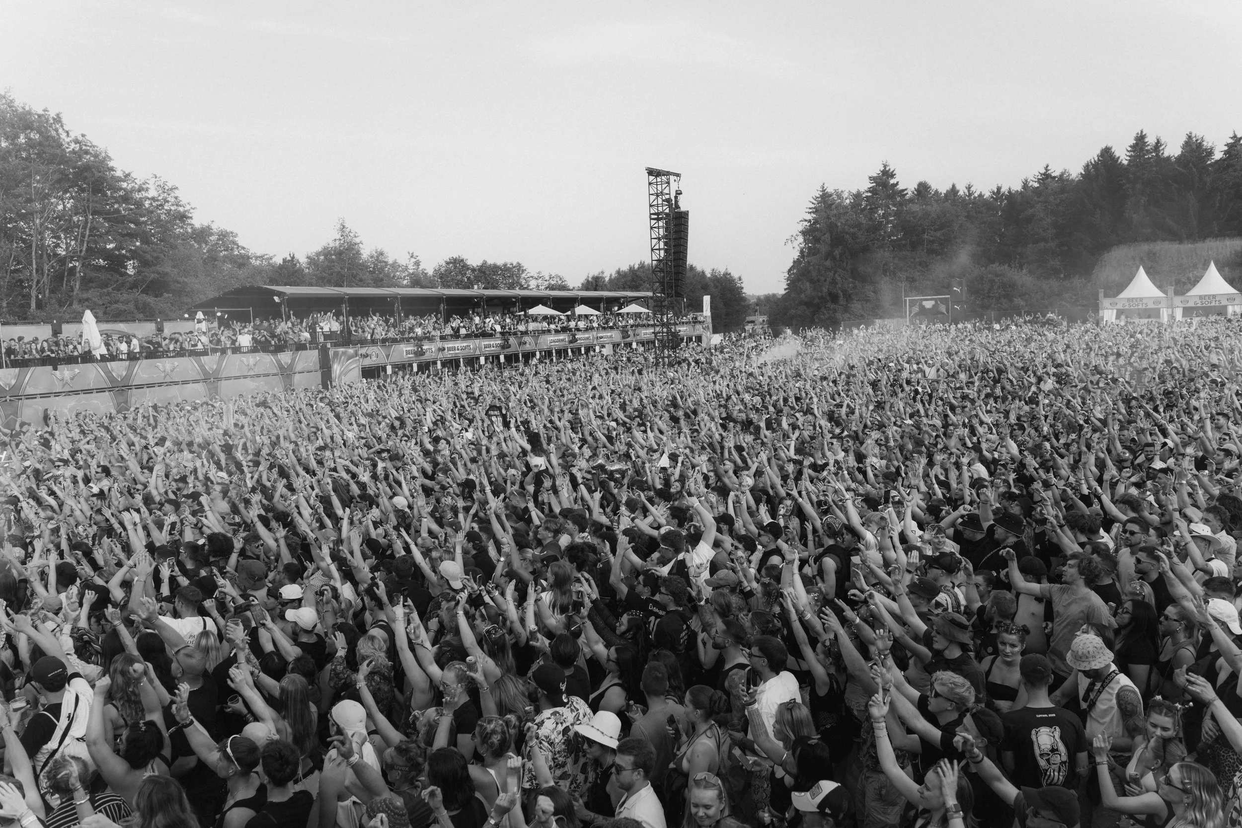 A large crowd of people at an outdoor music festival or concert, raising their hands and enjoying the event, with flags and tents visible in the background.