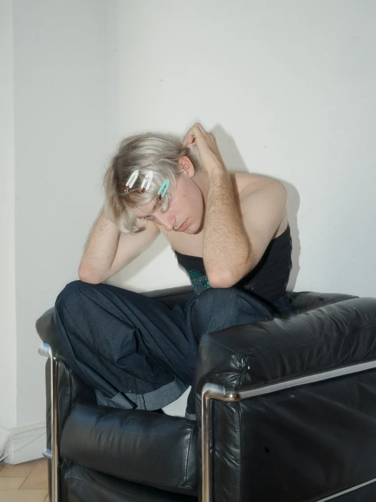Young person sitting on a black leather chair, leaning forward with their head in their hands, wearing a strapless top, with hair clips in their blonde hair, looking distressed.