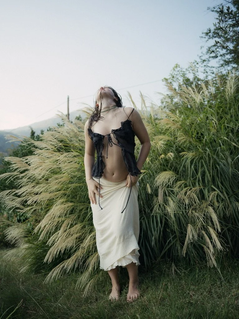 A woman wearing a black top and a long white skirt standing barefoot among tall grass.