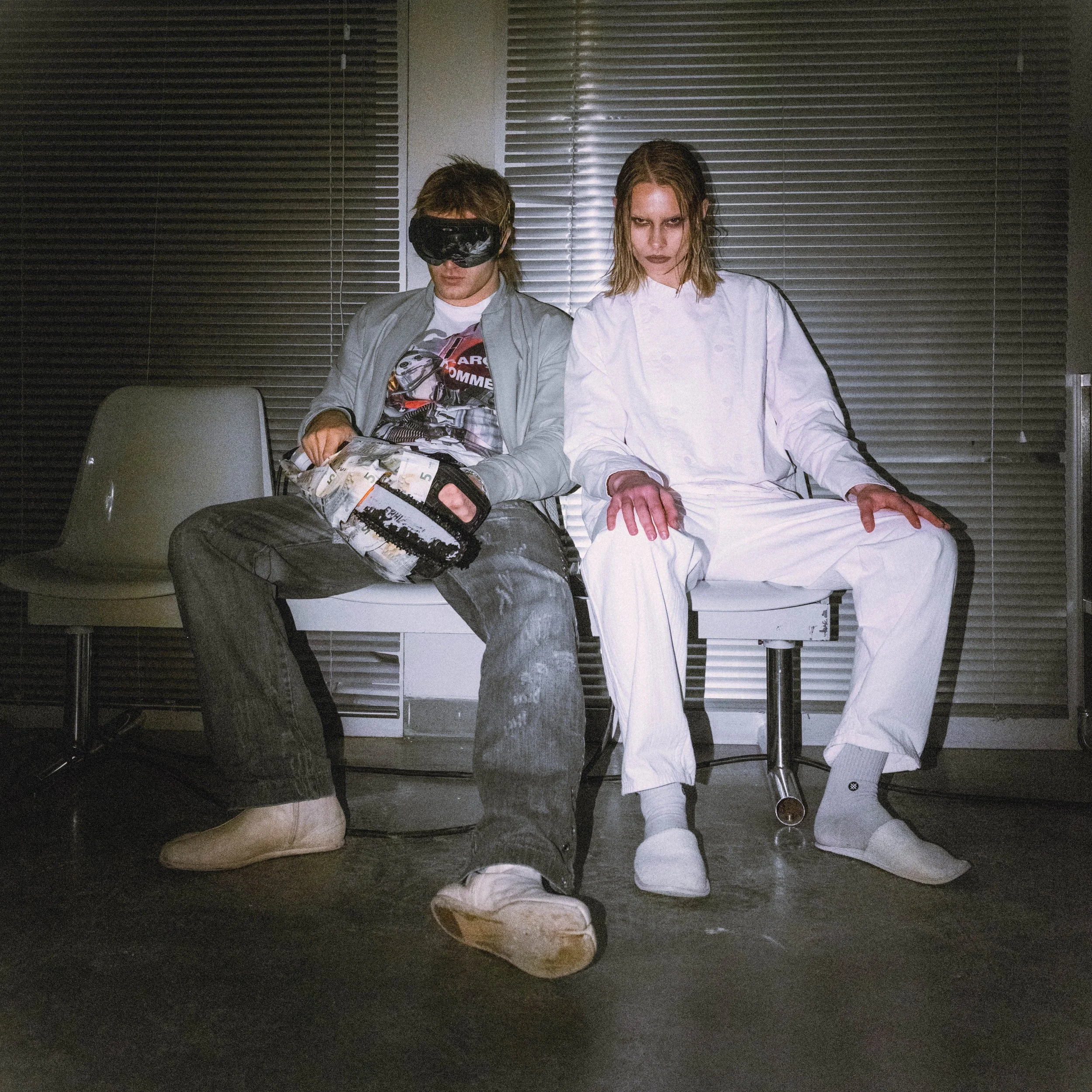 A young man with long hair wearing a gray jacket and a graphic t-shirt, and a young woman with long hair and dramatic makeup, dressed in a white chef's uniform, sit side by side against a wall with metal blinds. They are sitting on chairs, with the man's head slightly lowered and the woman's head slightly tilted, both appear to be looking downward. The surroundings suggest an indoor setting with artificial lighting.