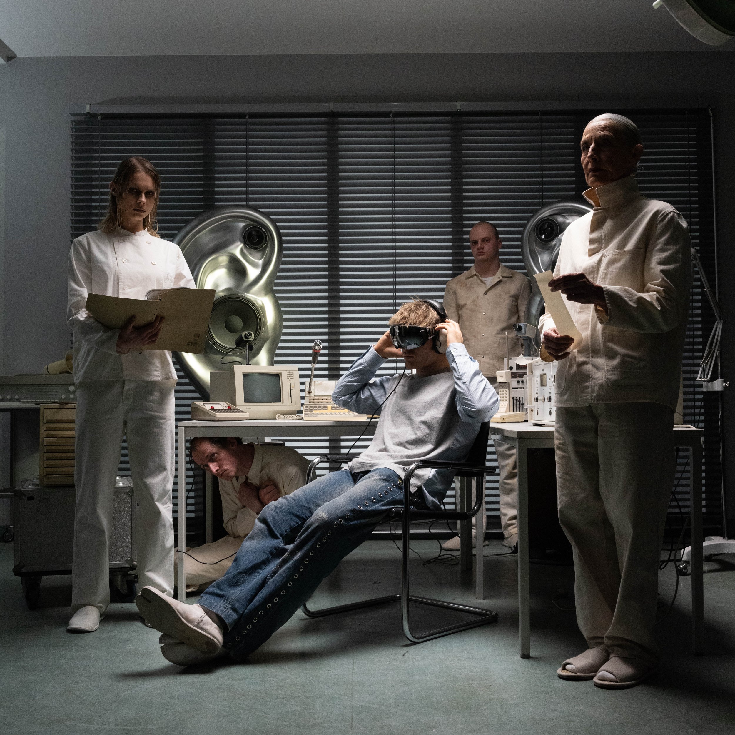 A group of people in a laboratory or industrial setting with dark blinds and large metallic speakers in the background, some wearing white coats or uniforms, some holding papers or using electronic devices, and one person sitting with their legs extended and crossed at the ankles.