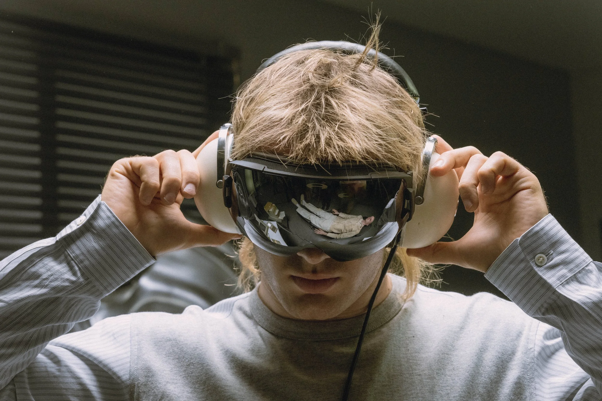 A person with blond hair is holding a virtual reality headset's straps with both hands on his head, looking down, wearing a light gray shirt, and in a room with dark ceilings and walls.