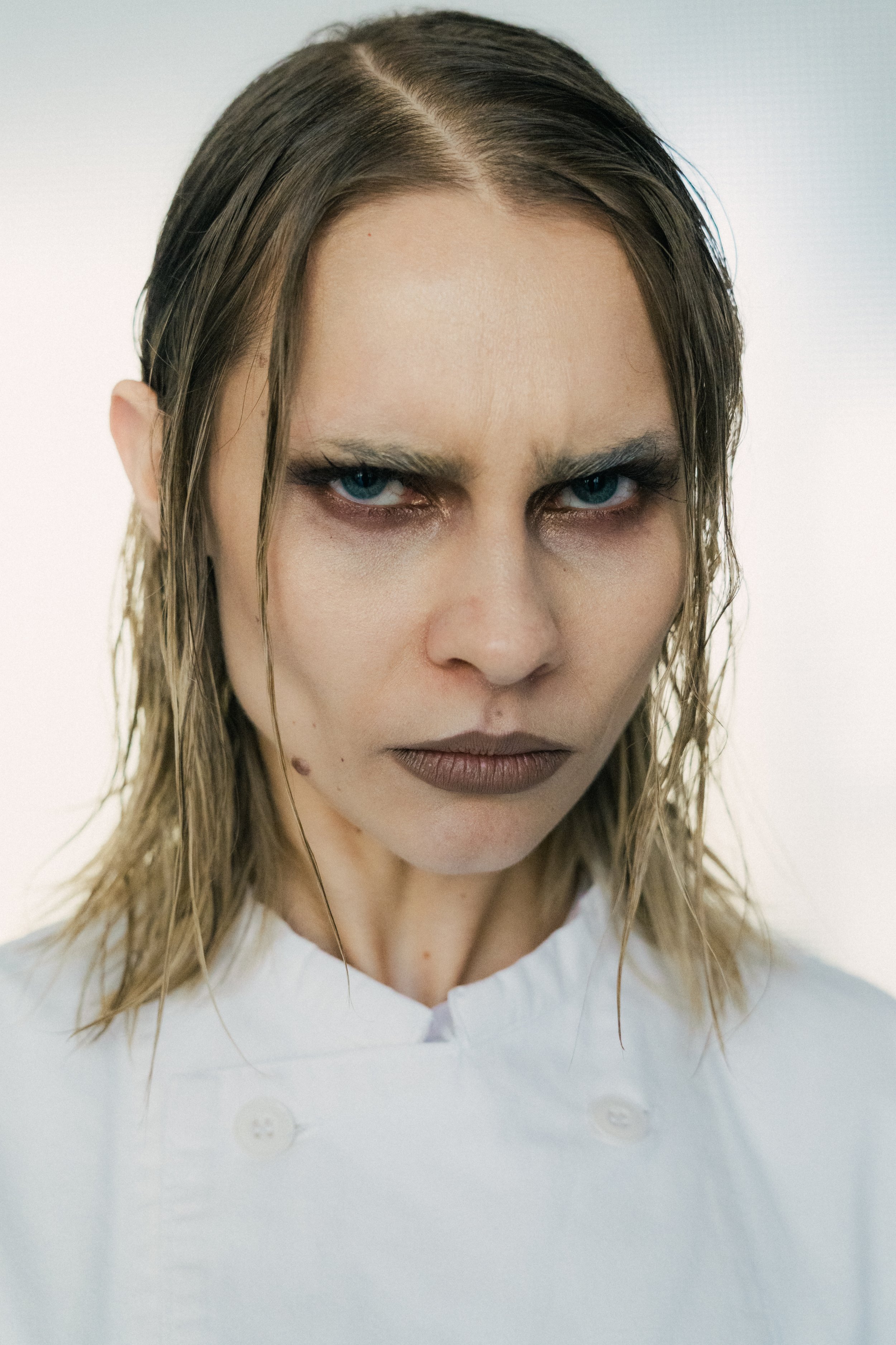 Close-up of a woman with wet hair, intense blue eyes, and a serious expression, wearing a white top, against a plain light background.