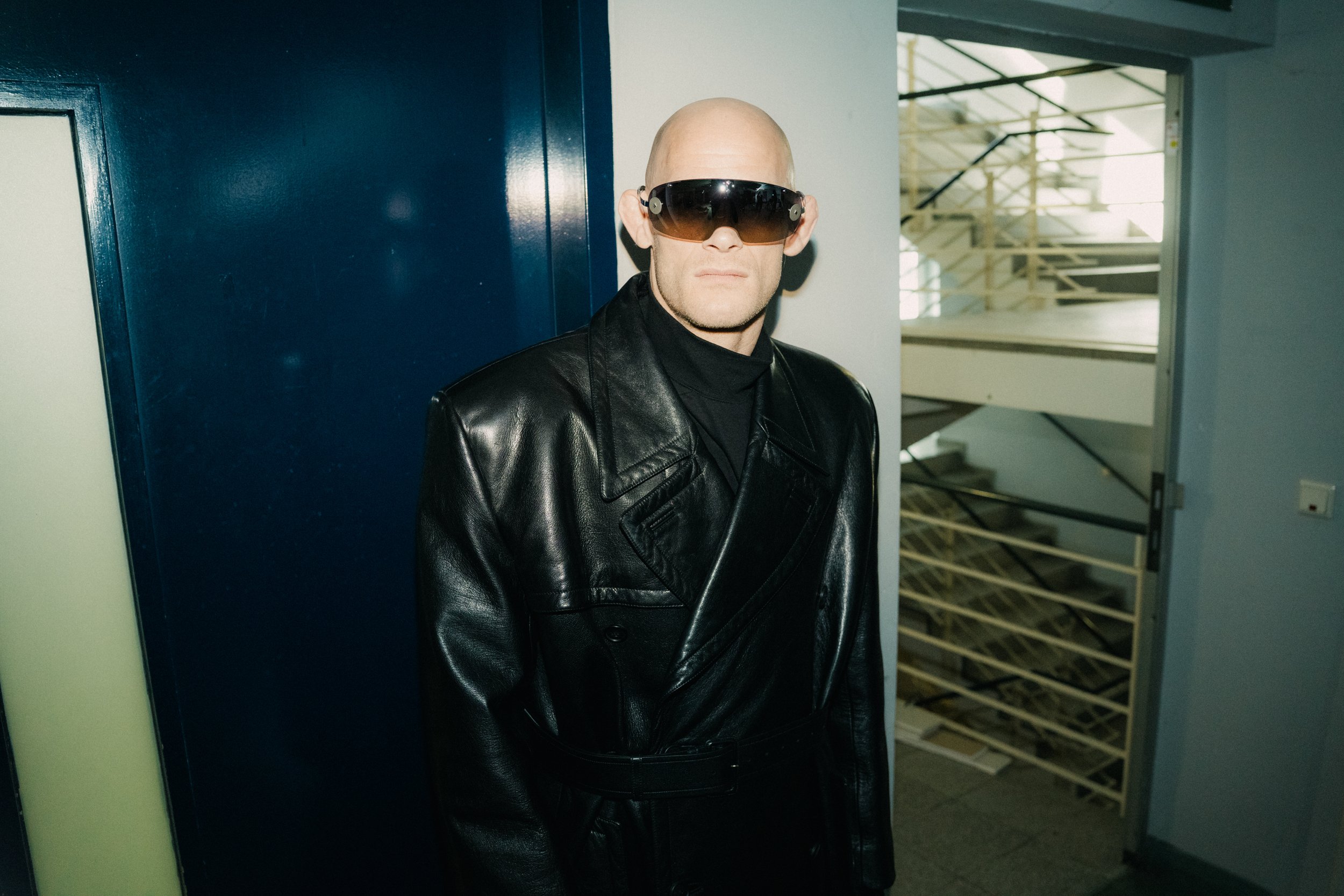 A bald man wearing black leather jacket and large sunglasses, standing in front of a staircase and interior wall.