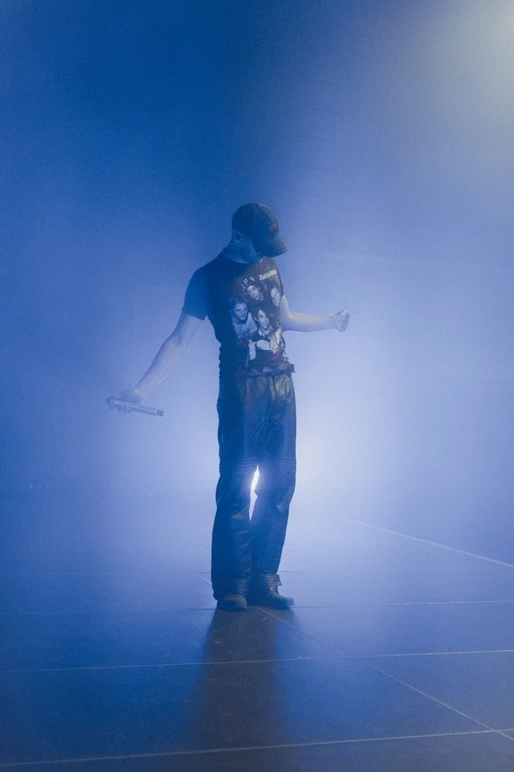 Person wearing a black cap, t-shirt with illustrated characters, and black pants, standing on stage with blue lighting, holding a microphone in one hand and looking down.