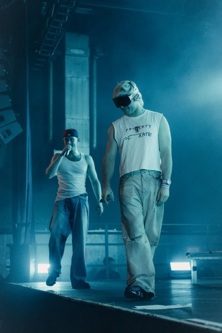 A man wearing a blindfold and casual clothes, holding a microphone, walking on stage during a performance or rehearsal, with a woman standing behind him in a similar casual outfit in a dimly lit, industrial-style venue.